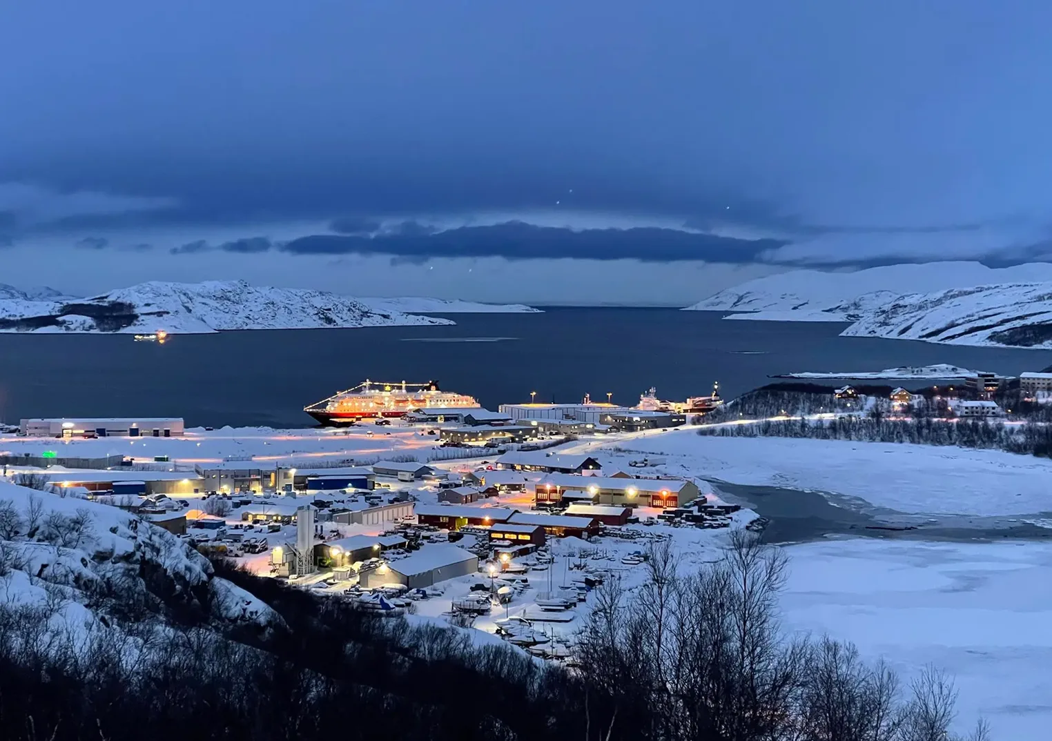 An aerial view of a snowy coastal town at dusk, with buildings lit up, a large ship docked, and snow-covered mountains surrounding a dark bay.
