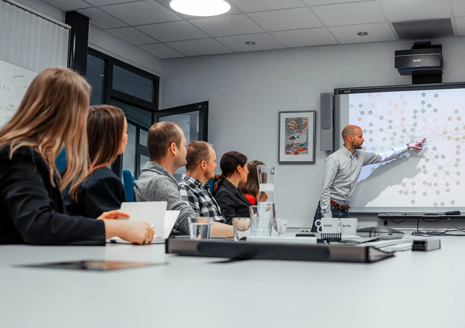 A man presents to a group in a modern meeting room, pointing at a network diagram displayed on a large interactive screen.