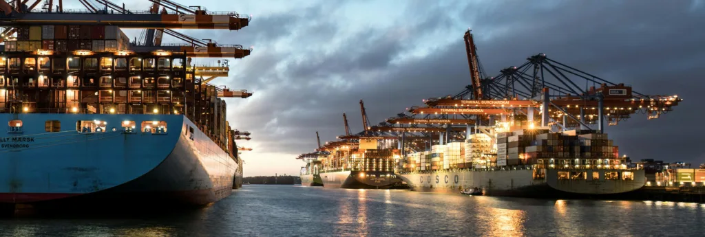 Large container ships docked at a bustling port at dusk, illuminated by numerous lights and cranes loading containers.