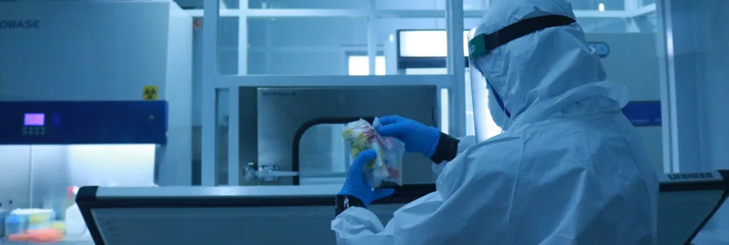 Scientist in a white hazmat suit and blue gloves handles a colorful sample in a sterile laboratory setting.