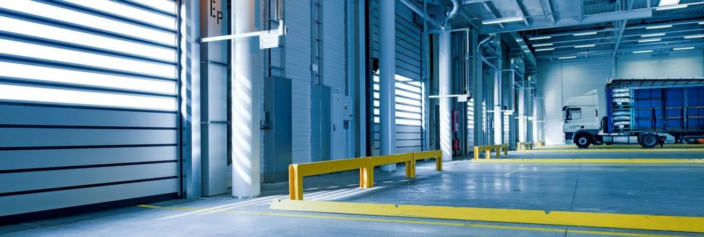 An industrial loading bay with a large white truck backed into a dock, surrounded by yellow safety barriers and markings.