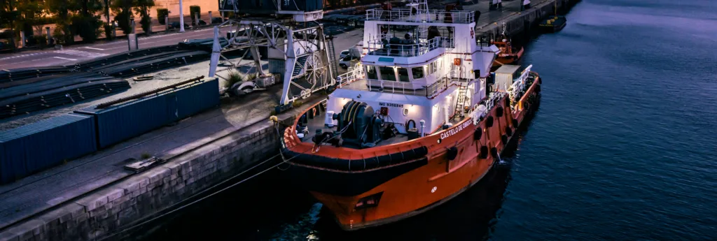 An orange and white tugboat docked at a dimly lit industrial port at dusk, with cranes and containers visible.