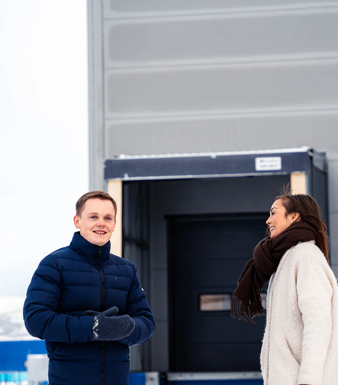 Two people, a man and a woman, stand outside a building in winter attire, with snow on the ground. The man smiles at the camera.