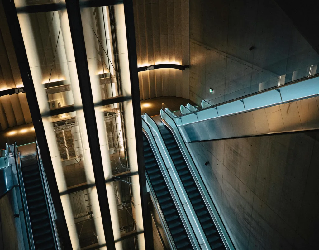 View of multiple escalators and a glass elevator shaft in a modern, brightly lit building or subway station.