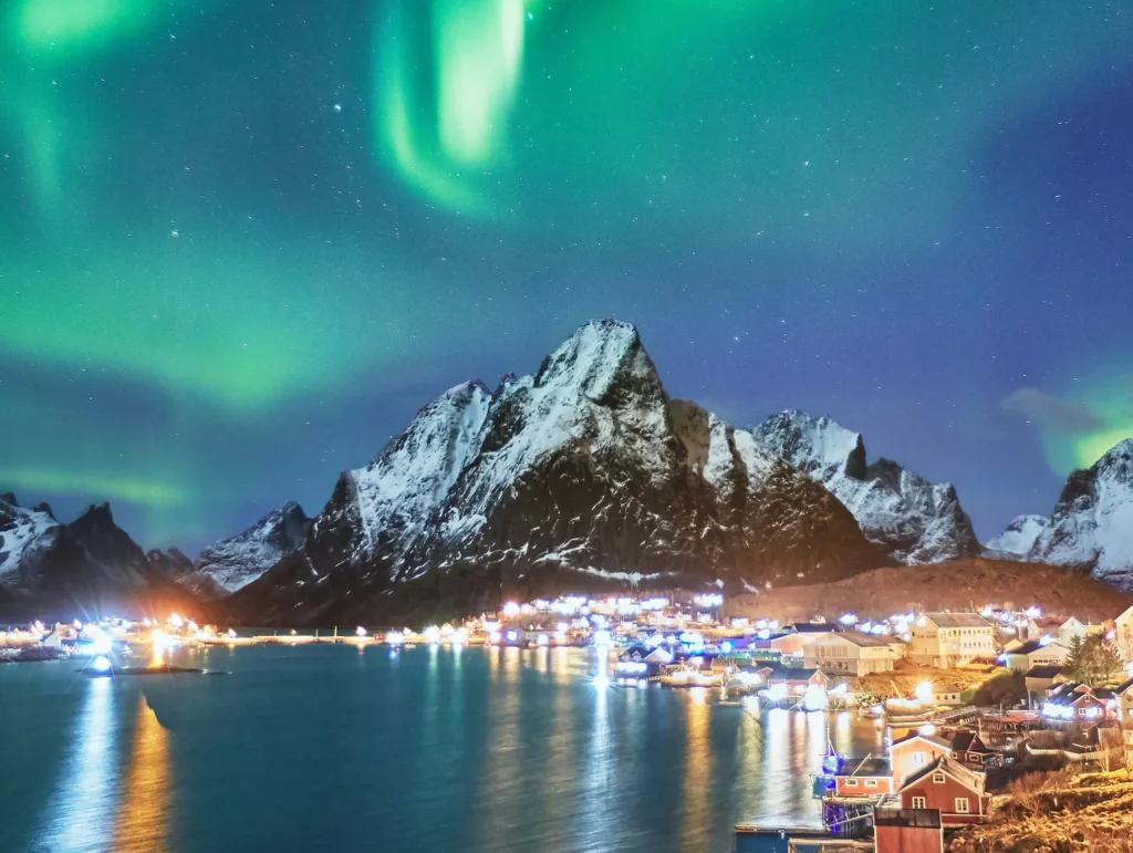 Scenic night landscape of a small village by a fjord, featuring illuminated buildings and their reflections, with large mountains under a starry sky.