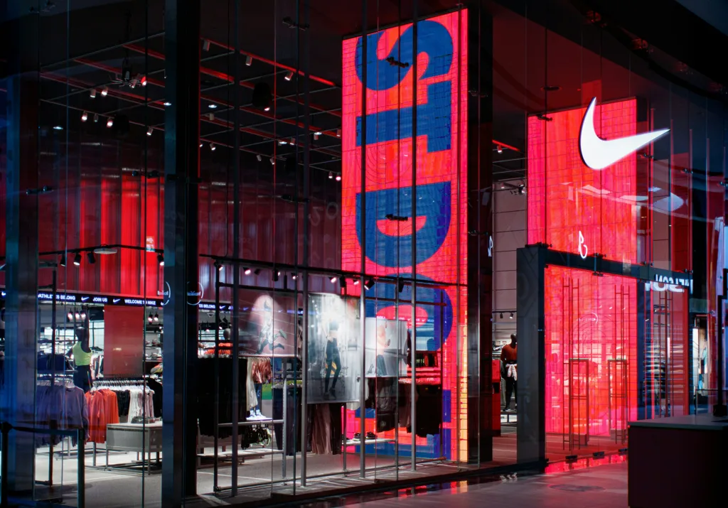 Exterior of a Nike store with large, vibrant red and blue digital displays showcasing branding and products. The store has a modern, illuminated glass facade.