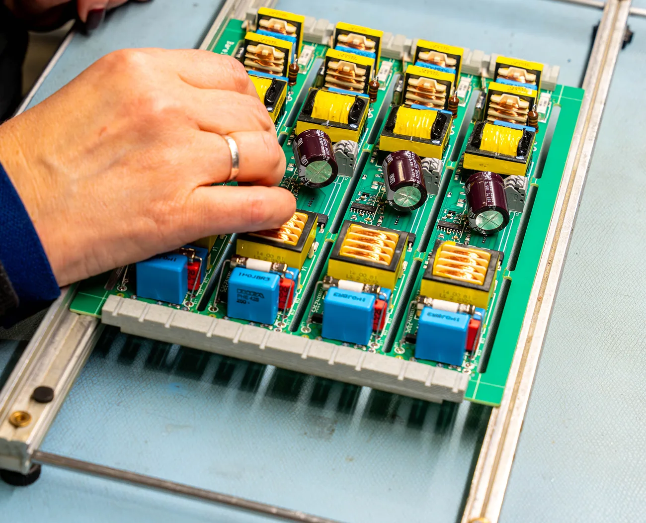A hand works on assembling or repairing a green circuit board with multiple yellow transformers and dark purple capacitors.