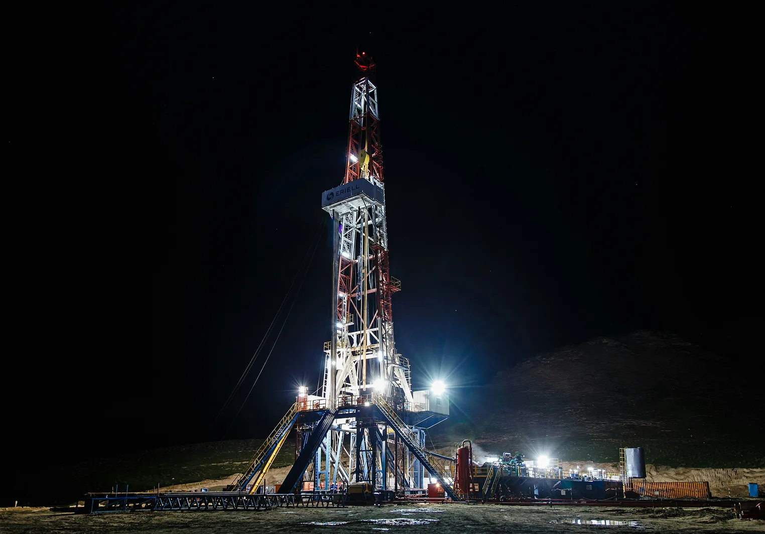 An illuminated oil rig, with a tall derrick structure and various industrial machinery, operates at night under a dark sky, highlighting its function.