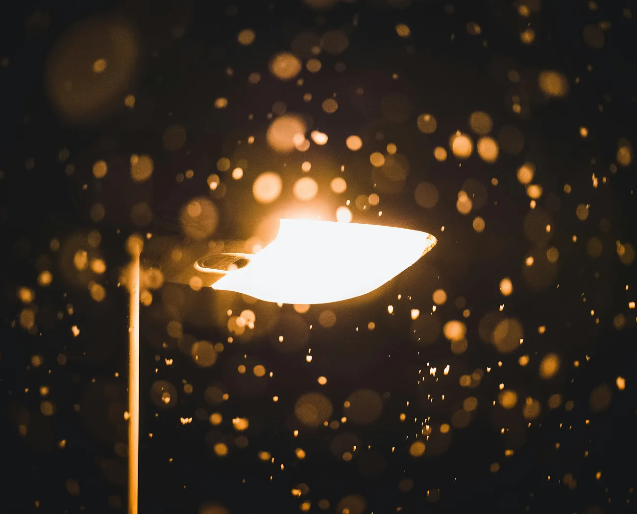 A bright street light illuminated at night, surrounded by numerous glowing bokeh lights from falling rain or snow.