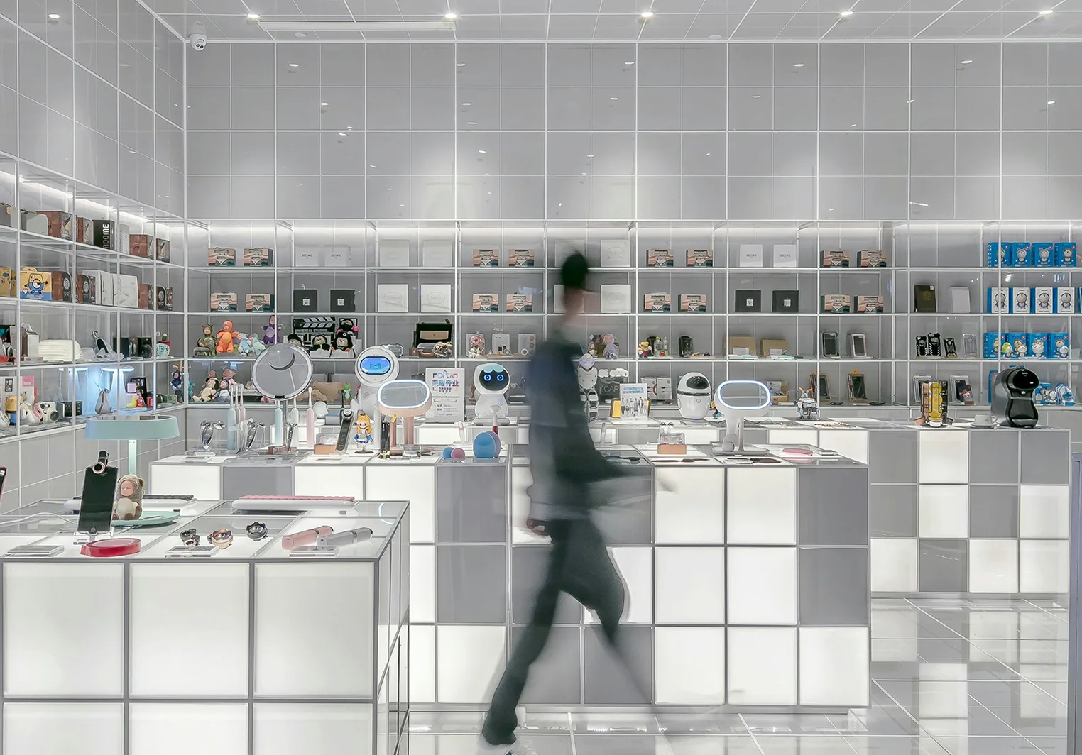 A brightly lit, modern electronics store featuring shelves filled with gadgets, robots, and packaged goods, with a blurry person walking by.