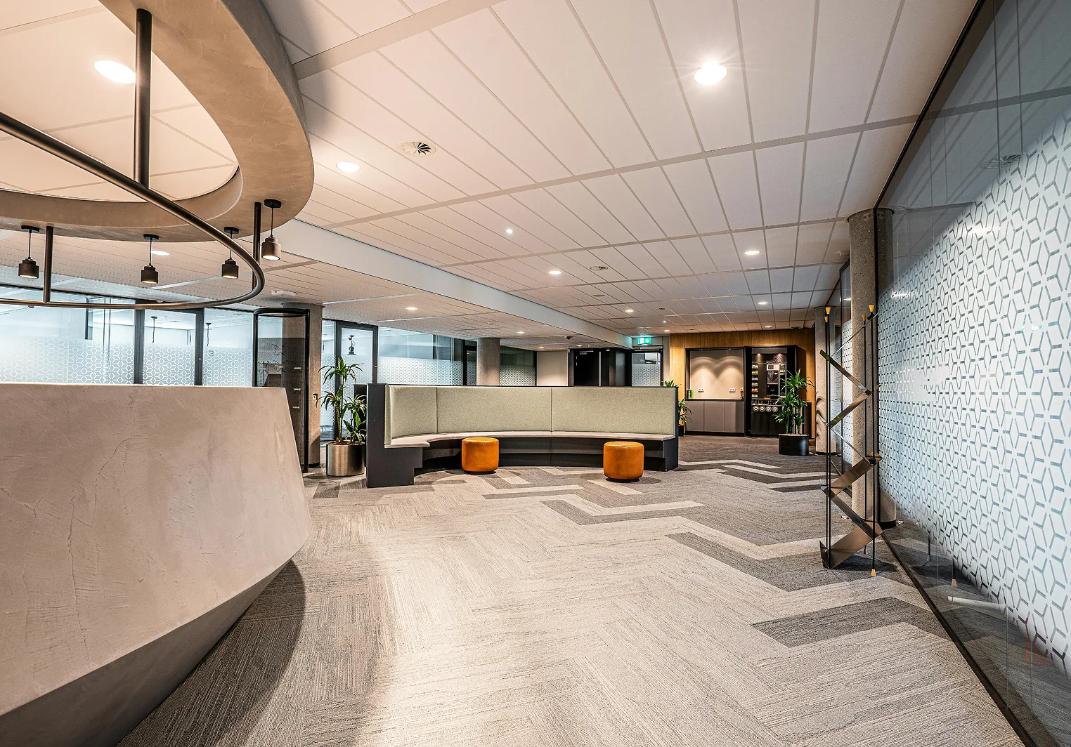 Modern office reception area with a curved desk, bench seating, patterned carpet, and glass walls.