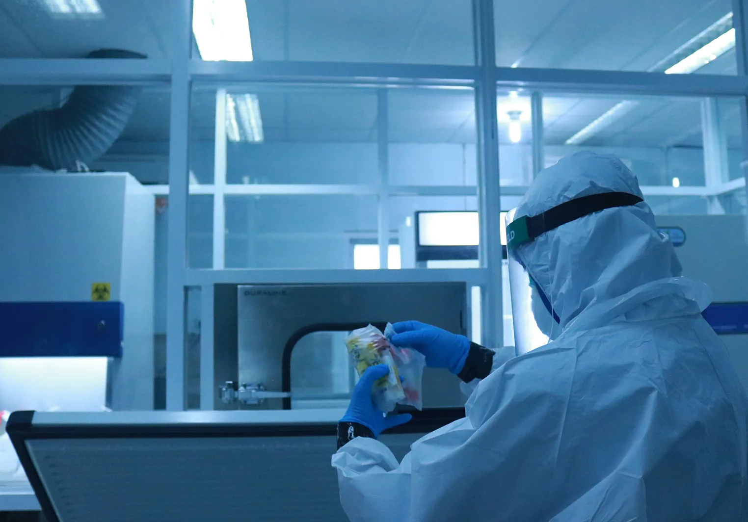 A person in a full white hazmat suit, blue gloves, and a face shield handles a clear bag with colorful items in a laboratory.