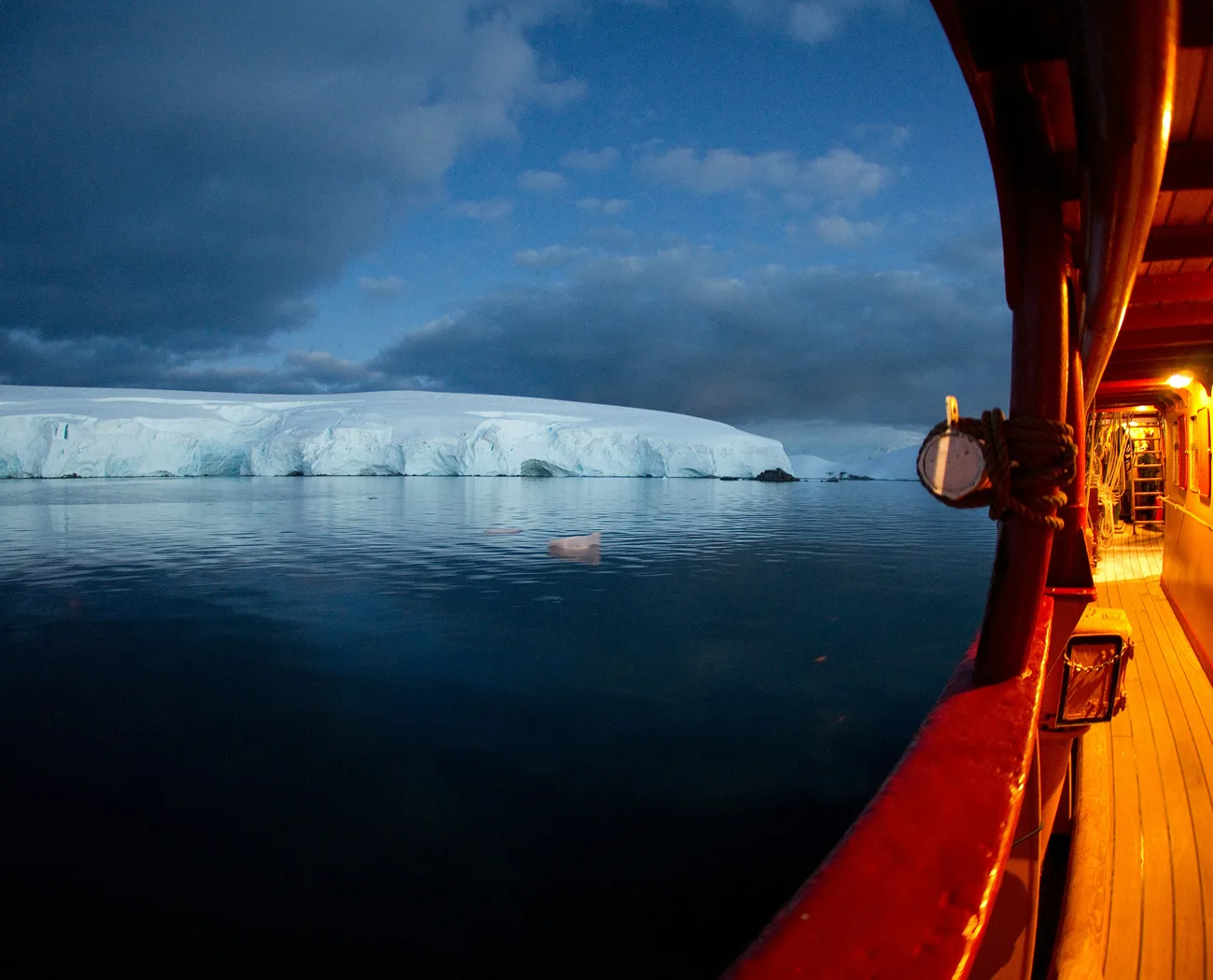 A ship's deck lit by warm lights overlooks calm, dark water with a large ice sheet and clouds at twilight.