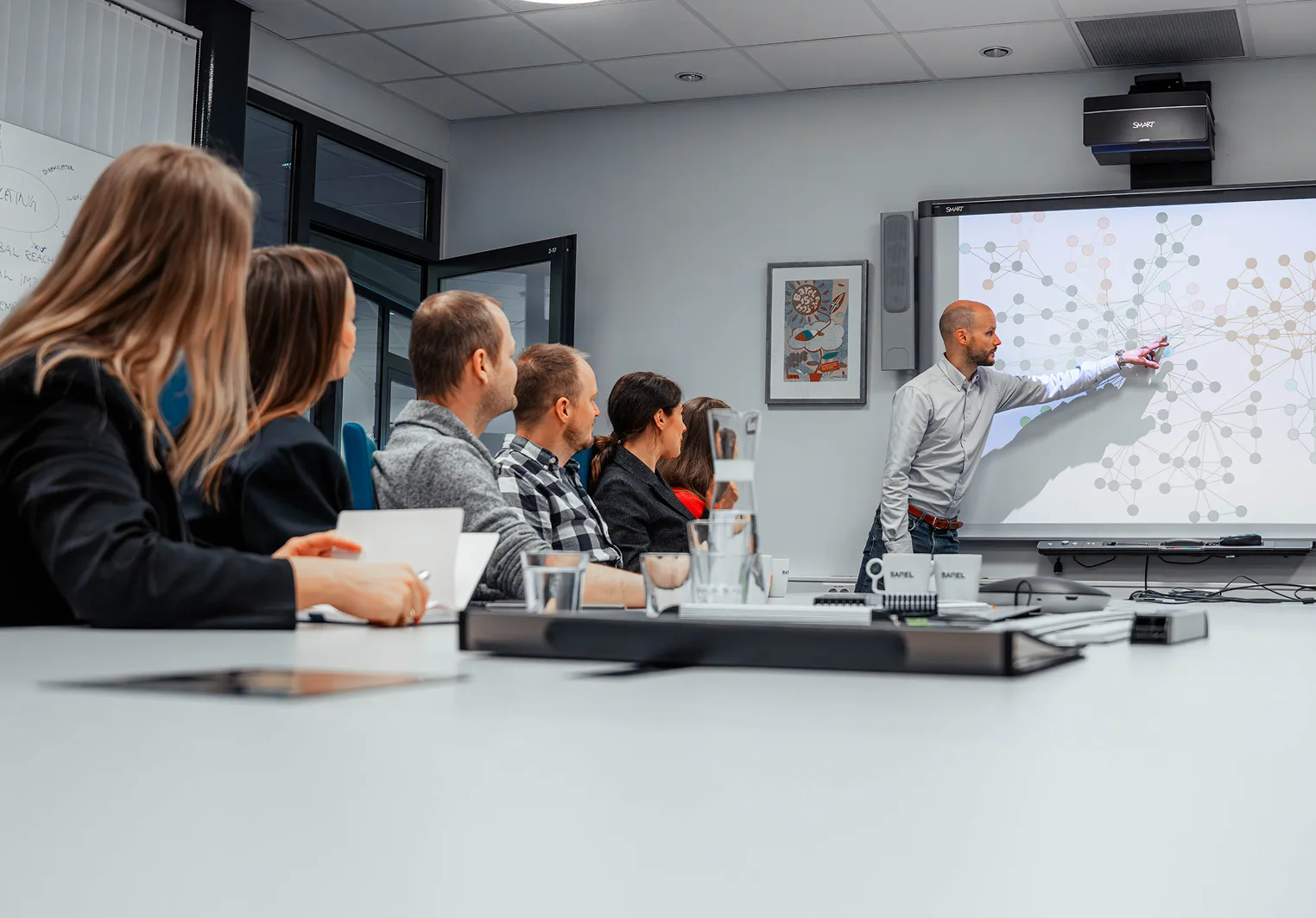 A group of professionals attends a business meeting, with a presenter actively explaining a diagram on a large interactive screen.