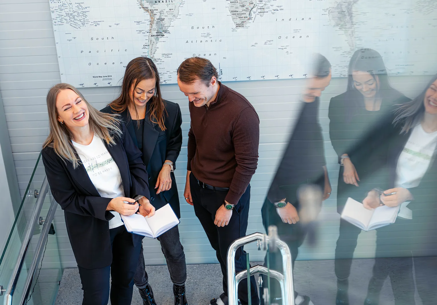 Four colleagues laugh and engage in a discussion near a world map, some holding notebooks.