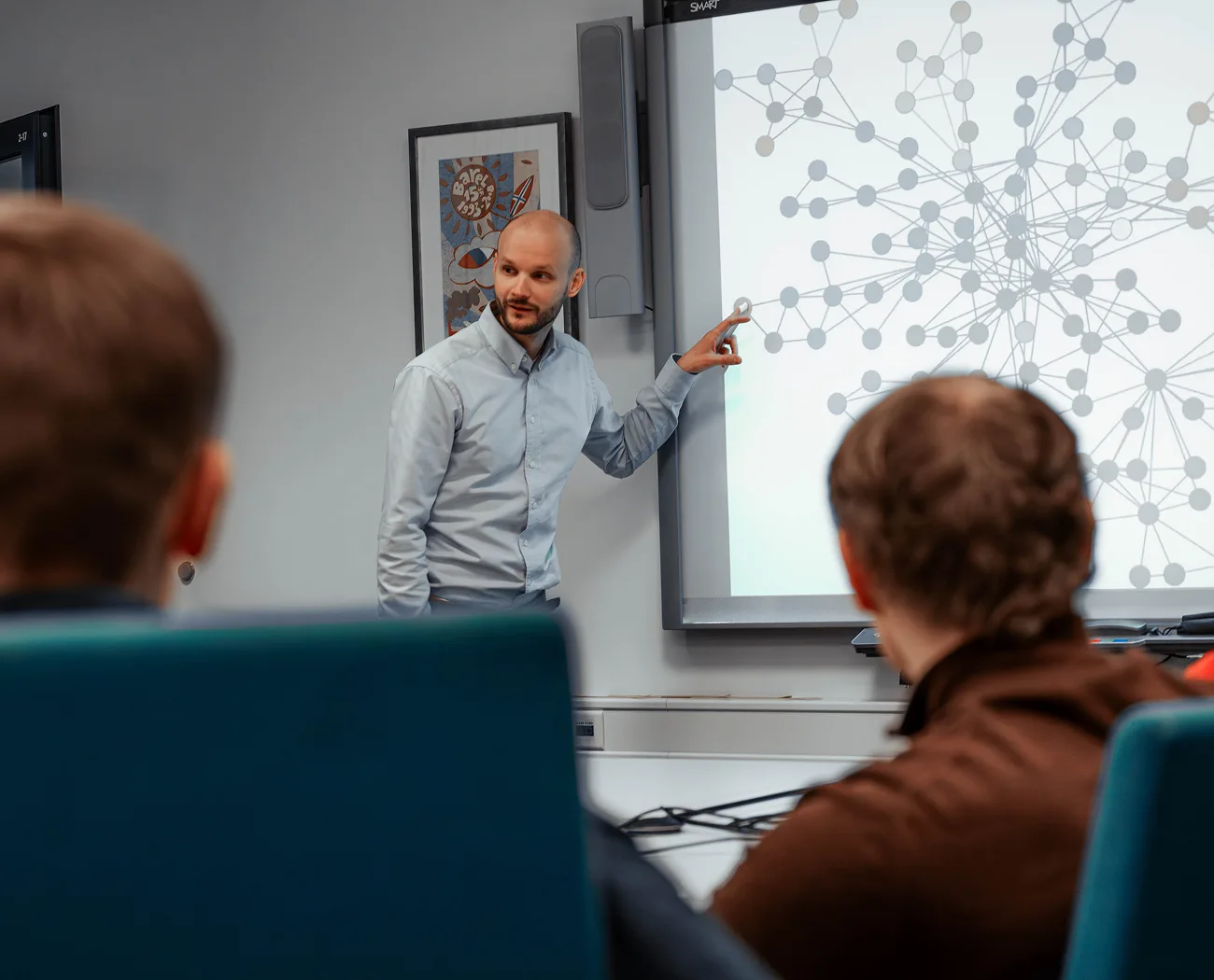 A man in a light blue shirt points to a network graph on a large screen during a presentation to an audience.