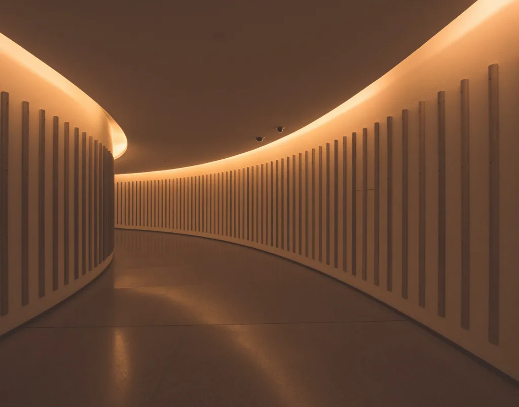 A modern, curved hallway illuminated by warm orange lights along the ceiling, with vertical textured panels on the white walls.