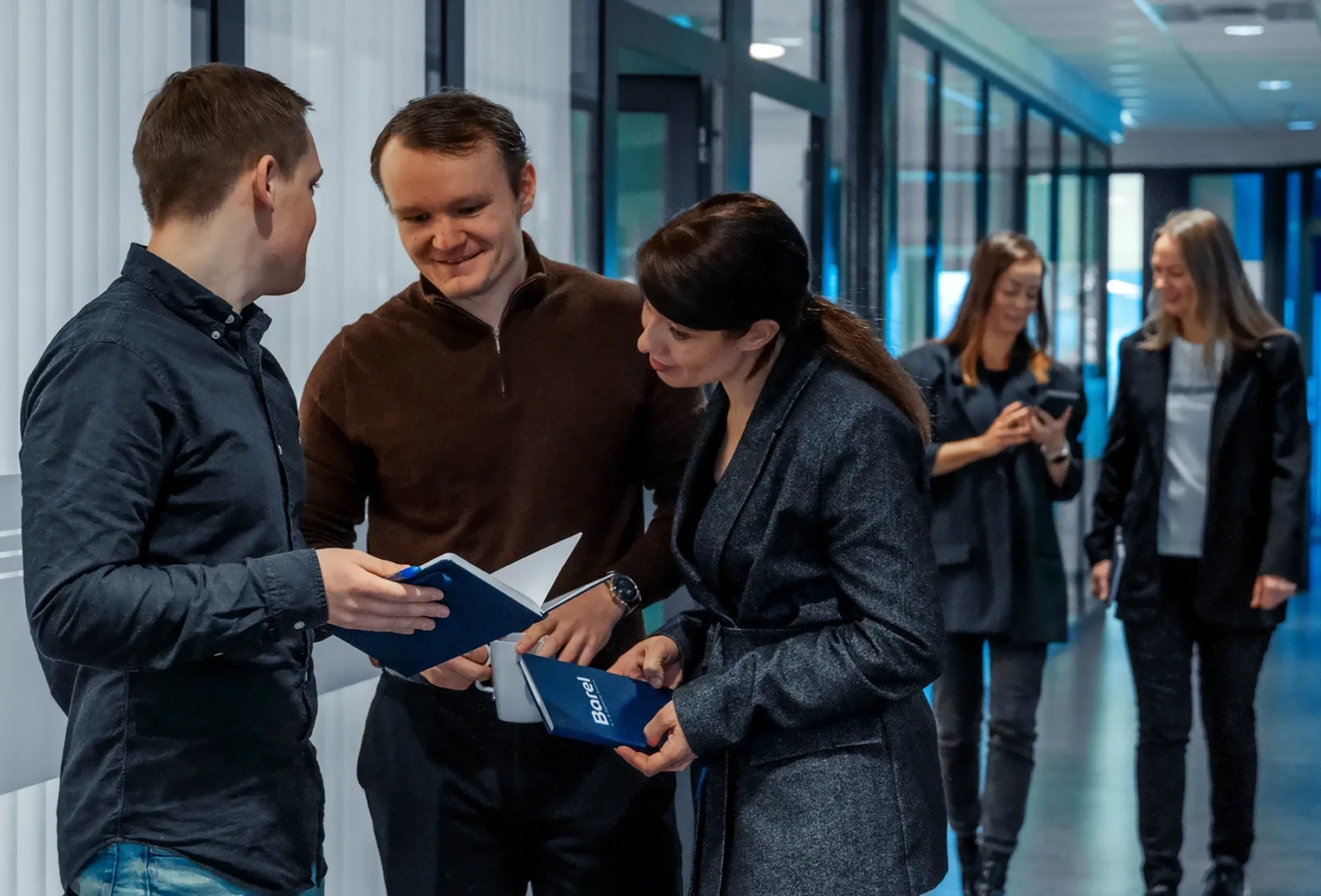 Three colleagues review documents in an office hallway, with two more people walking in the background.