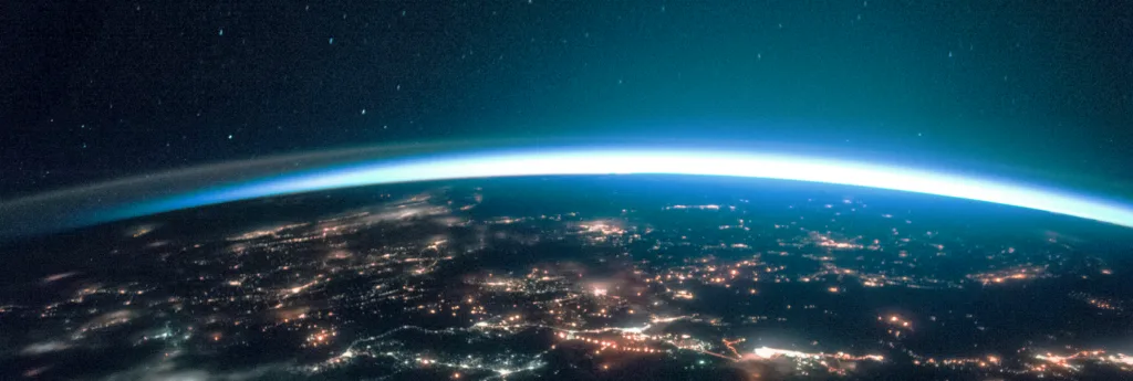 View of Earth from space at night, showing city lights and the thin glowing atmosphere against a dark sky.