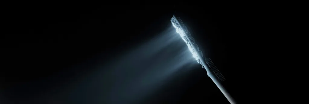 A long, slender light fixture emitting bright, cool-toned light diagonally against a dark background with a subtle dot pattern.