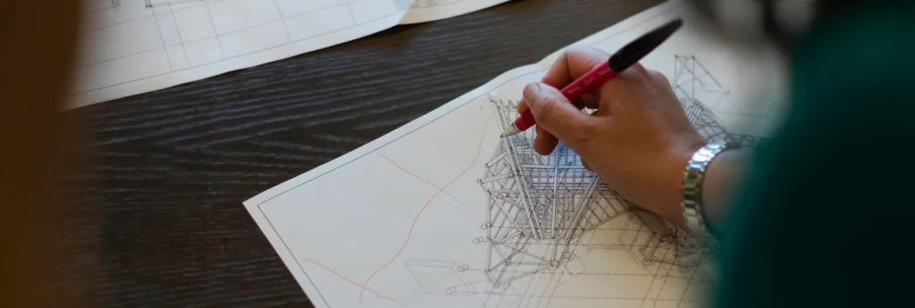 A person's hand with a watch, actively sketching on a detailed architectural plan or engineering drawing.
