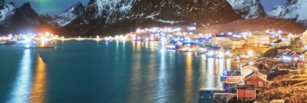 Scenic night landscape of a small village by a fjord, featuring illuminated buildings and their reflections, with large mountains under a starry sky.