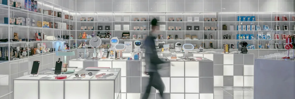 Interior view of a modern retail store displaying various electronic gadgets and consumer goods on white shelves and counters.