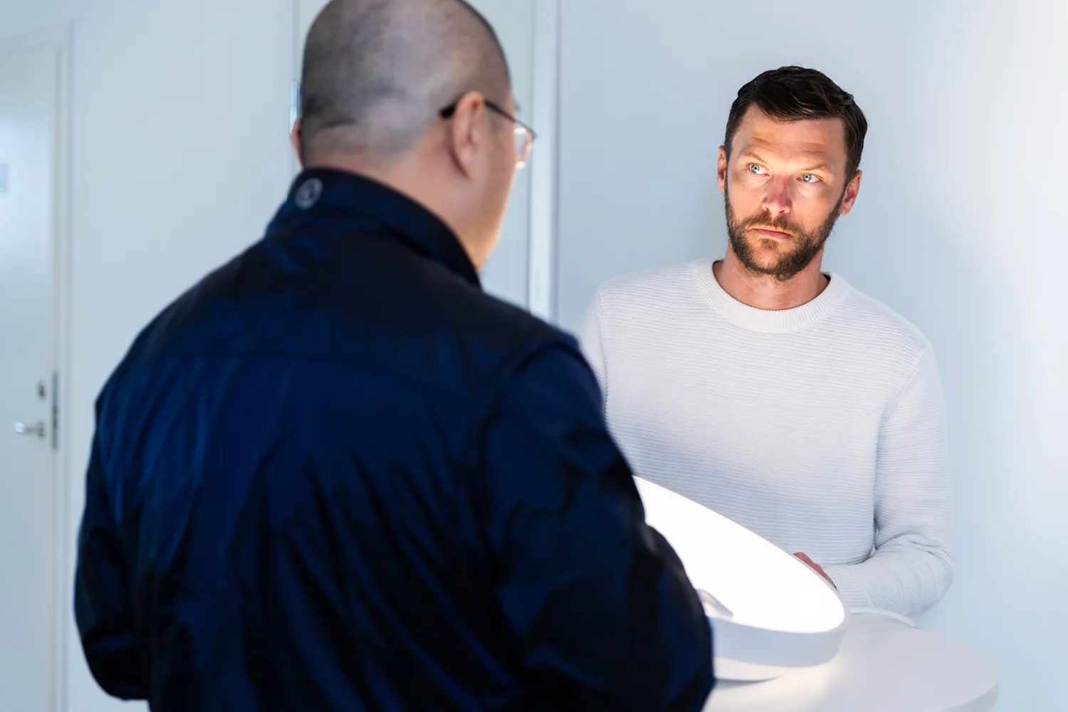 Two men discussing, with one facing away and the other looking intently, a glowing circular light on a white table between them.
