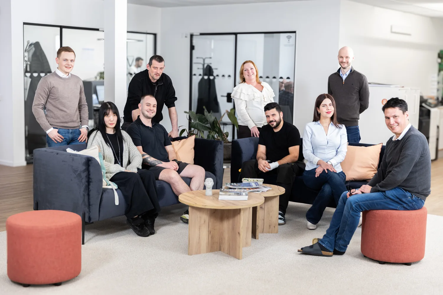 A diverse group of ten employees smiles at the camera in a modern office lounge area. Some are seated on sofas, others stand, showcasing a collaborative team.
