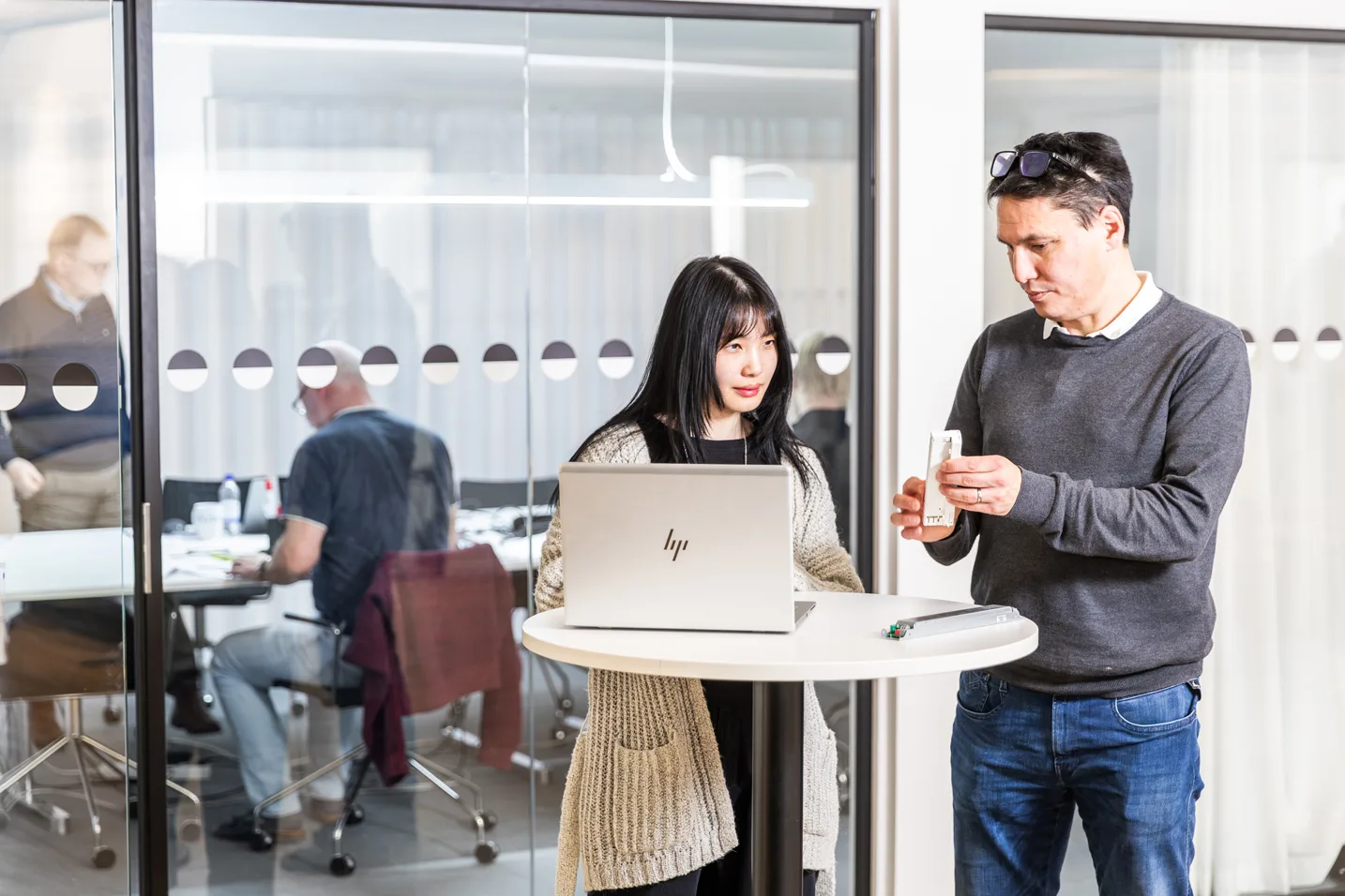 Two colleagues, a woman and a man, standing at a table with a laptop and electronic devices in an office setting.