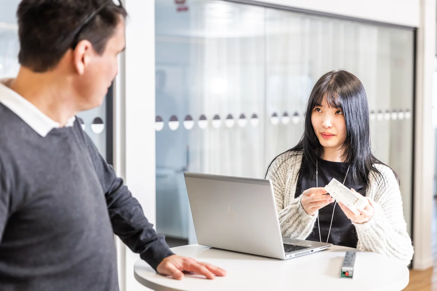 A woman holding a white device discusses with a man at a standing desk with a laptop.