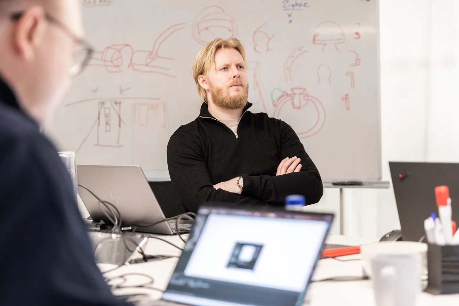 Man with beard and black sweater listening attentively in a meeting, with a whiteboard of diagrams behind him.