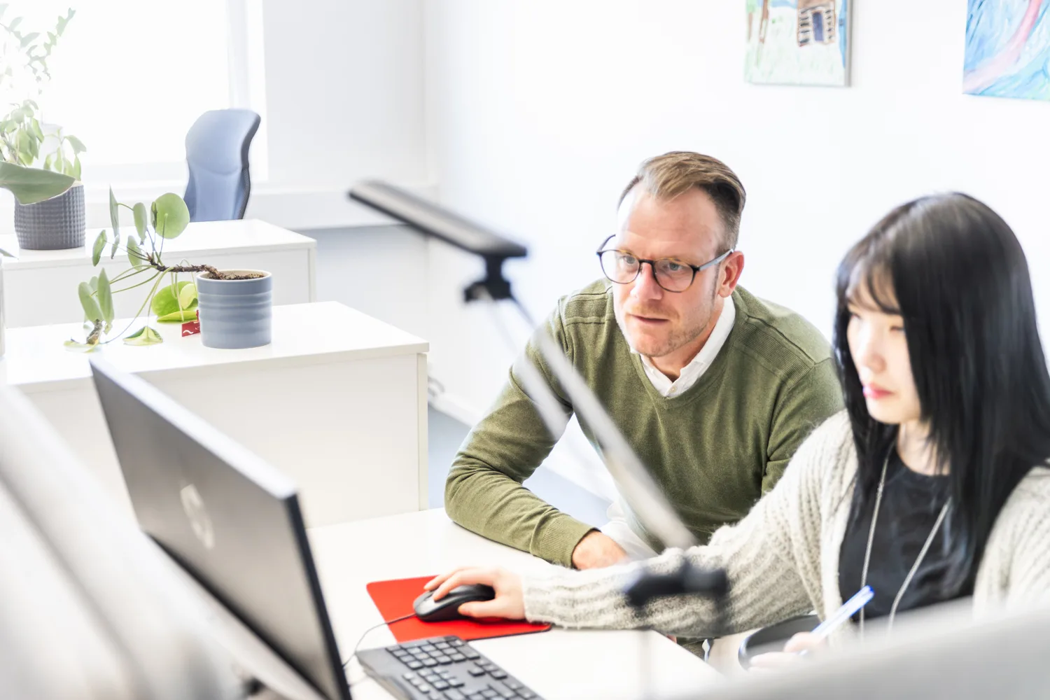 A man in glasses and a woman with dark hair work together at a computer in a bright office environment.