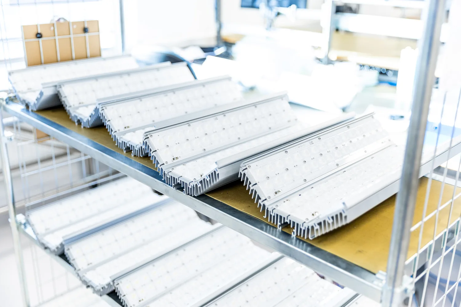 Rows of uninstalled LED light fixtures with heat sinks on a metal shelf in a factory or warehouse setting.