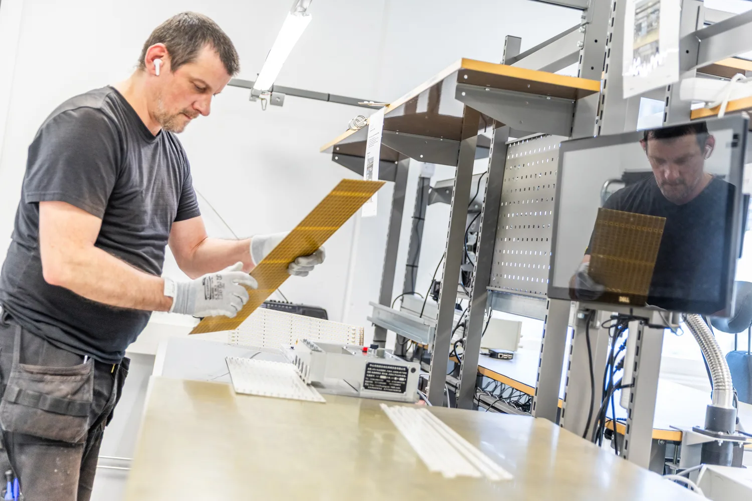 An employee in a manufacturing facility examines a golden circuit board with a grid pattern, wearing protective gloves. A tool belt with "aluwave" is visible.