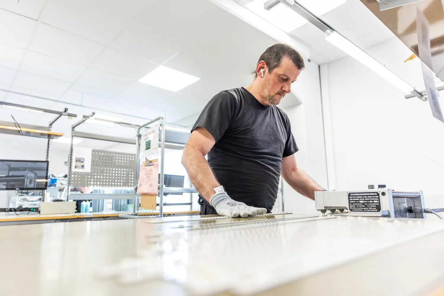 Worker in a manufacturing setting carefully uses a tool on a flat surface, focusing on a precise task.