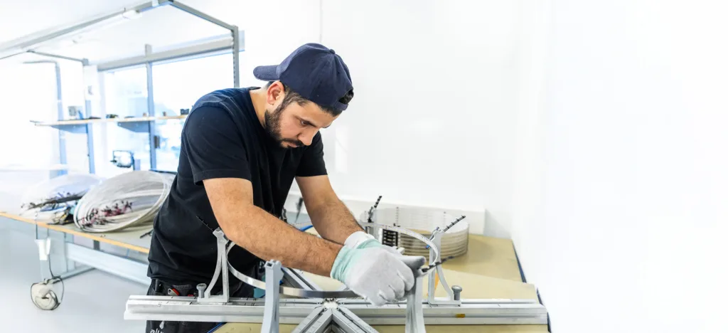 Man in a cap and gloves assembling circular components on a workbench in a workshop.