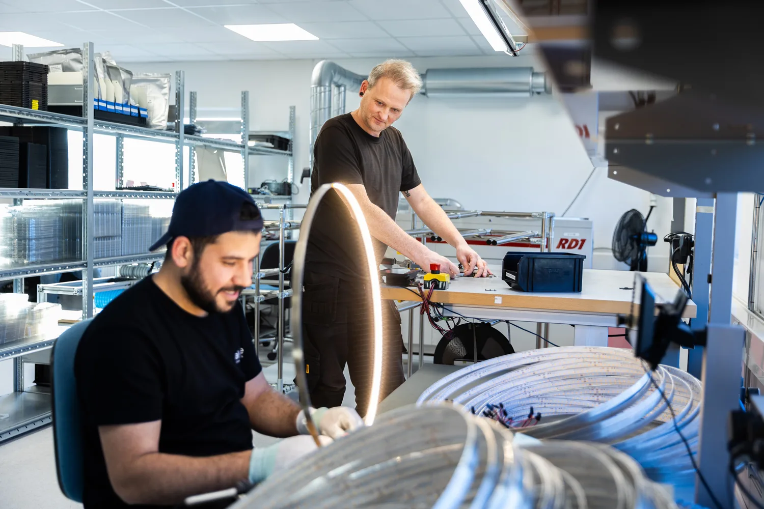 Two men in a workshop assembling wires and components, with shelves and equipment in the background.
