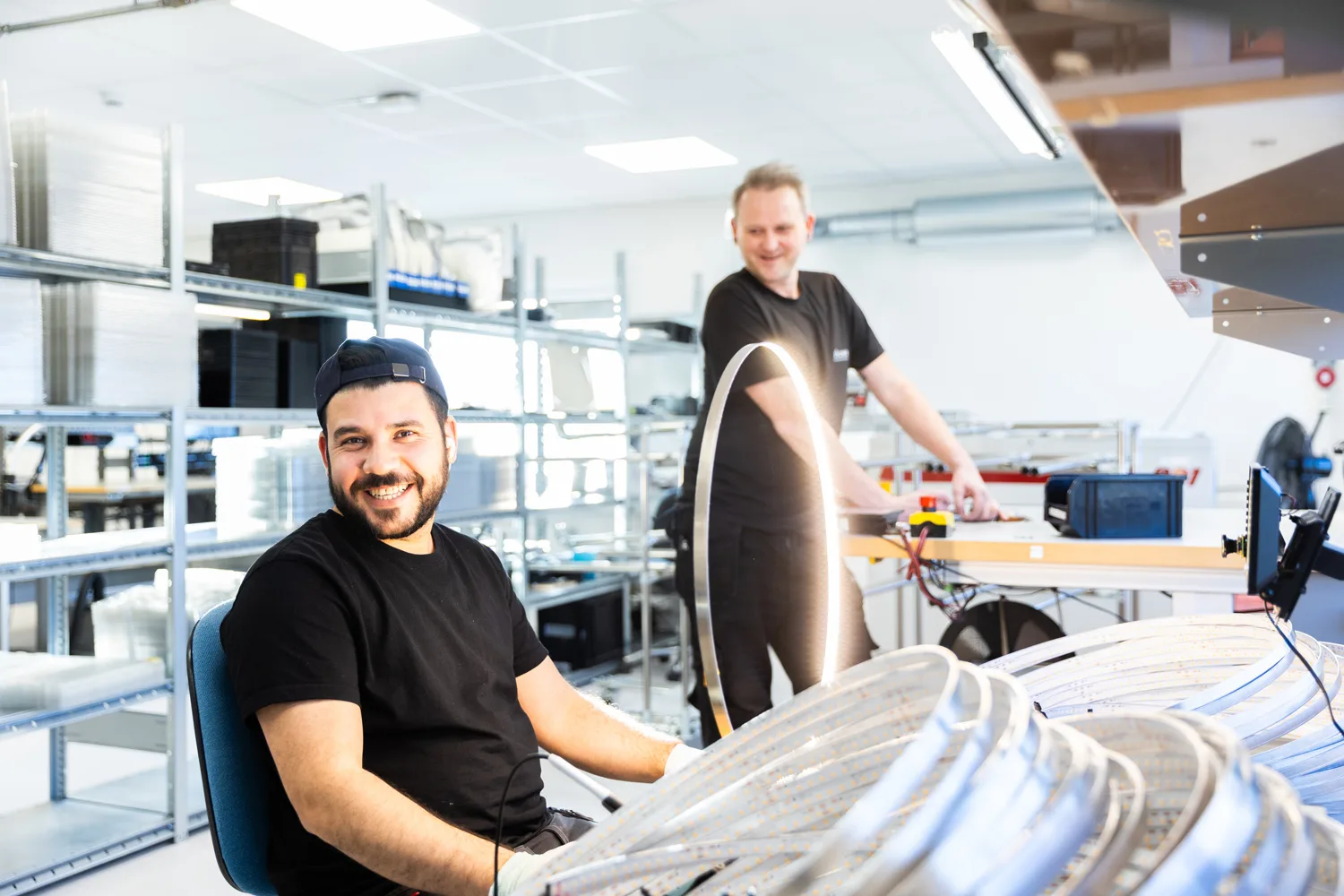 Two smiling men in a workshop, one seated looking at the camera, the other working at a bench.