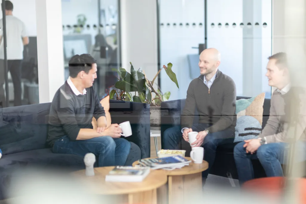 Three colleagues review documents in an office hallway, with two more people walking in the background.